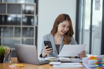 Fototapeta premium Accounting asian woman use smartphone and laptop at office desk in office, Accounting businesswoman online working concept.