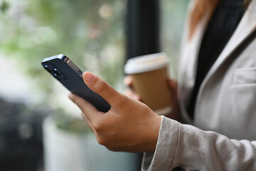 Unrecognizable young businessman holding paper cup and using mobile phone near office window