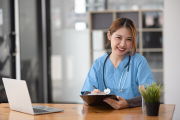 Asian female doctor working with a patient clipboard and digital laptop sitting at desk in...