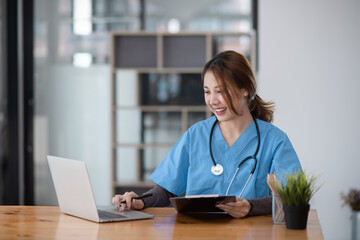 Asian female doctor working with a patient clipboard and digital laptop sitting at desk in hospital, Medicine and healthcare concept.