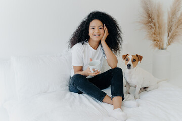 Relaxed Black woman with curly hair enjoying coffee on bed with her Jack Russell Terrier