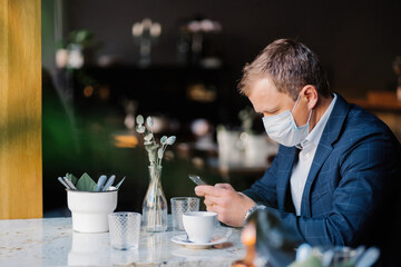 Businessman in a suit using smartphone at café with mask on, maintaining social distance