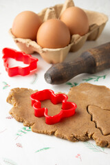 Top view of gingerbread cookie dough with red Christmas molds, tree and doll, on white paper with rolling pin and eggs, vertical