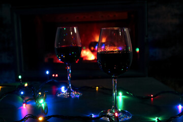 Two red wine glasses and lights decoration on burning fireplace background. Wine by the fireplace.