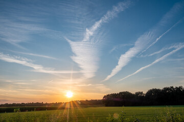 The image captures a majestic sunset over a verdant field, where the sun kisses the horizon, sending rays of light across the sky. The dramatic clouds are etched into the blue expanse, forming