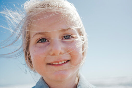 A girl on the beach with a joyful expression and wind-swept hair, her eyes squinting slightly in the bright sunlight, captures the essence of beachside bliss.