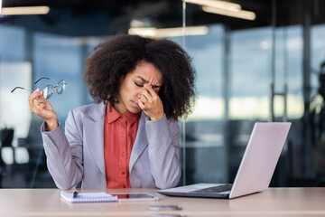 Overworked tired business woman has dizziness and pain in eyes, woman took off glasses and rubs bridge of nose, works long time at laptop in middle of office, accountant in jacket. - obrazy, fototapety, plakaty
