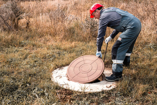 A worker in a hard hat lifts a manhole cover on a septic well. Inspection and maintenance of sewerage systems in rural areas