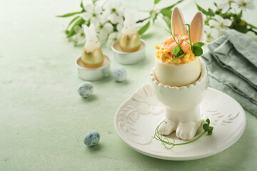 Fototapeta na wymiar Stuffed or deviled eggs with yolk, shrimp, pea microgreens with paprika in rabbit-shaped stand for easter table decorate fresh cherry or apple blossoms on light background. Traditional dish for Easter