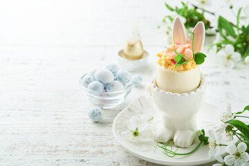 Stuffed or deviled eggs with yolk, shrimp, pea microgreens with paprika in rabbit-shaped stand for easter table decorate fresh cherry or apple blossoms on light background. Traditional dish for Easter