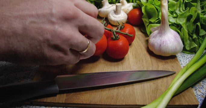  Background close-up of cherry tomatoes lying on a board next to a knife, onion, champignons, garlic and lettuce.Human hand taking and putting it.