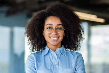 Close up of young beautiful woman with curly hair and glasses, businesswoman at workplace looking at camera, satisfied female worker in shirt smiling at workplace inside office