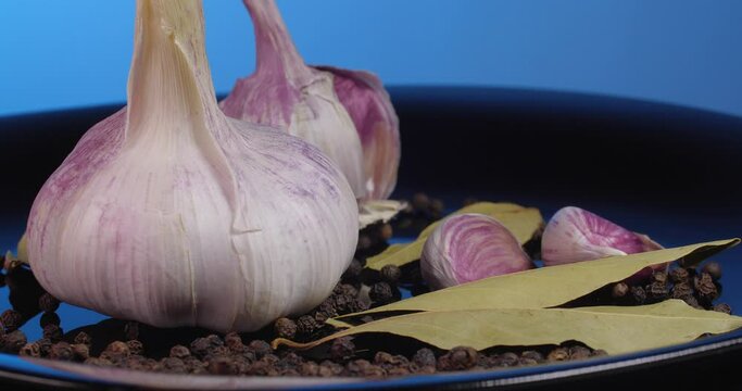 Close up garlic cloves with black peppercorns and  bay leaf rotate on a black plate on a blue background.