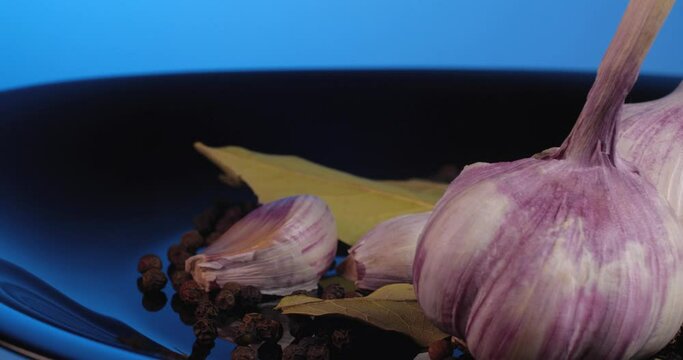 Close up garlic cloves with black peppercorns and  bay leaf rotate on a black plate on a blue background.