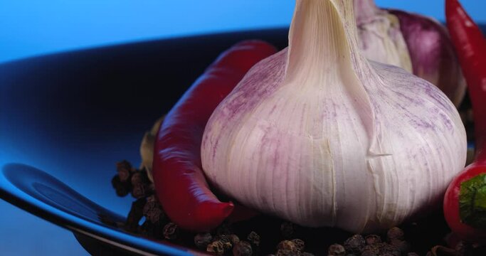 Close up garlic cloves with black peppercorns, red chilly pepper and  bay leaf rotate on a black plate on a blue background.