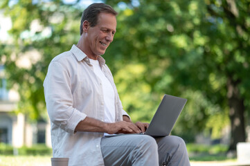 Middle aged freelancer programmer man sitting on bench working online on laptop in city park.
