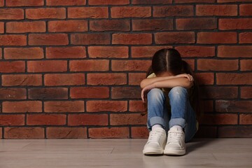 Child abuse. Upset little girl sitting on floor near brick wall indoors, space for text