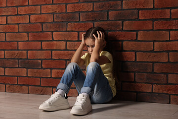 Child abuse. Upset little girl sitting on floor near brick wall indoors