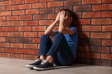 Child abuse. Upset boy sitting on floor near brick wall indoors, space for text