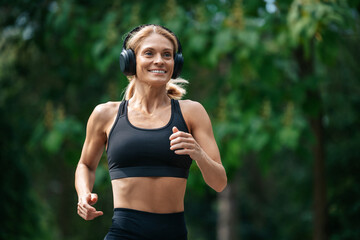 Athletic strong blonde runner woman in headphones jogging in park.