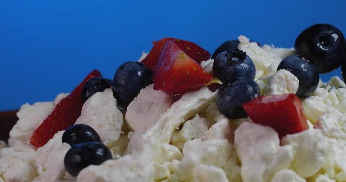 Extreme close up human hamd putting strawberries and blueberries on  cottage cheese rotate on a brown plate on a blue background.