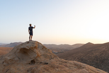 Rear View of Person Standing on a Rock Taking a Picture with Phone Against a Mountain Background