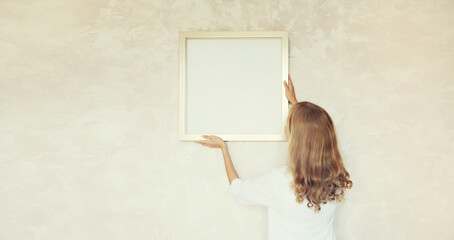 Young woman decorating interior, hanging blank a photo frame mockup on white wall in new home