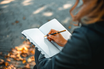 Young woman writing in a diary in an autumn park. Closeup image of a woman writing on a blank notebook. Woman working outdoors. - obrazy, fototapety, plakaty