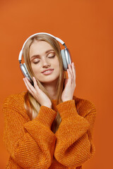 blonde woman in knitted sweater and headphones smiling with closed eyes on orange, winter leisure