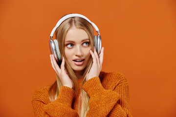 amazed blonde woman in bright knitted sweater listening music in headphones on orange, enjoyment