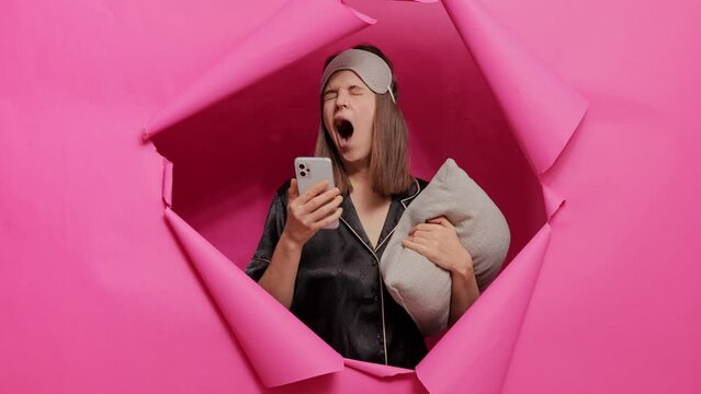 Sleepy woman wearing sleeping mask and pajama posing in paper hole of pink wall with pillow in hands using mobile phone after waking up browsing internet pages in the morning.