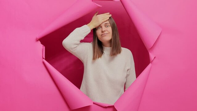 Worried sad brown haired woman wearing sweater posing through hole of pink paper wall making facepalm gesture bad memory forgot something important.
