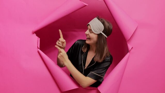 Smiling cheerful lady braking through pink paper torn wearing pajama and sleeping mask pointing away at copy space advertisement area indicating aside with happy face.