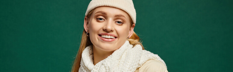 trendy woman in white knitted scarf and beanie hat smiling at camera on green, winter style banner