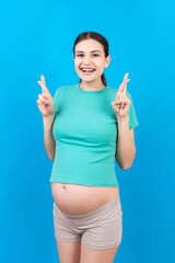 Fototapeta premium Cheerful young pregnant woman holding fingers crossed, waiting pregnancy isolated on colored background in studio. lifestyle pregnancy concept