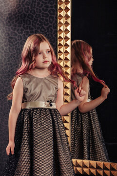 Portrait of fashionable cover child girl in stylish dress posing at large mirror, looking away. Adorable lovely kid lady stand in living dark room inside. Children emotion concept. Copy ad text space