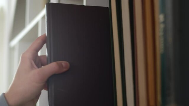 Close-up of a hand carefully taking a book out of big range staying in row in a shelf of a white bookcase at home. Concept of curiosity and thirst for knowledge