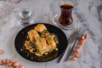 Traditional famous Turkish dessert baklava, pistachios and tea glass in black plate on marble...