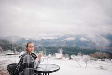 Woman sitting and holding cup and drinking hot coffee on winter holiday. Female drink tea and relaxing on cabin terrace with mountain view.  Girl in a blanket in winter on a hill near forest.