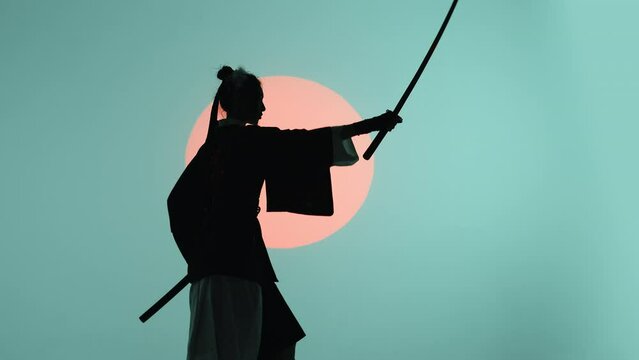 Silhouette of a young asian woman in a traditional kimono militantly removing the katana from the sheath and making its swing in the studio with a turquoise blue green background and red sun