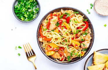 Stir fry egg noodles with chicken slices, pineapple, red paprika, chives, soy sauce and sesame seeds in ceramic bowl. Asian cuisine dish. White table background, top view