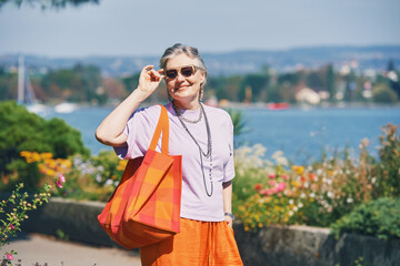 Outdoor fashion portrait of middle age 50 - 55 year old womn wearing colorful outfit, posing by the lake, travel and vacation lifestyle, image taken in Morges, Vaud, Switzerland