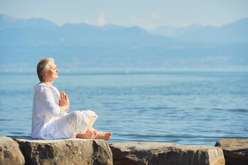 Outdoor portrait of happy mature woman meditating by the lake or sea, wearing white clothes,...