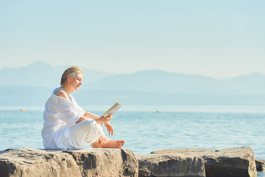 Outdoor portrait of happy mature woman reading book by the lake or sea, summer vacation - Powered by Adobe