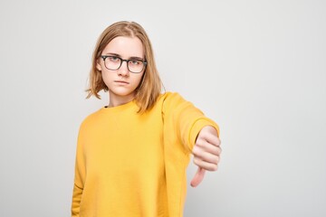Portrait of young student girl in glasses and yellow cardigan showing thumbs down isolated on white...
