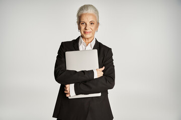 smiling middle aged businesswoman in formal wear standing and holding laptop isolated on grey