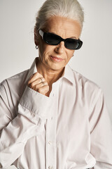 fashionable middle aged woman in classy white shirt and sunglasses posing on grey background