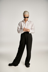 full length of stylish middle aged woman in white shirt, black pants and sunglasses posing on grey