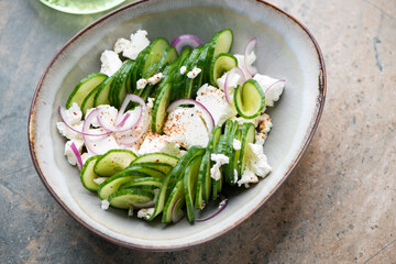 Salad with fresh cucumber, feta cheese and red onion in a grey bowl, horizontal shot on a beige granite background