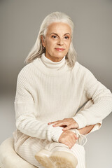 attractive grey haired middle aged woman in white winter outfit sitting on pouf on grey background